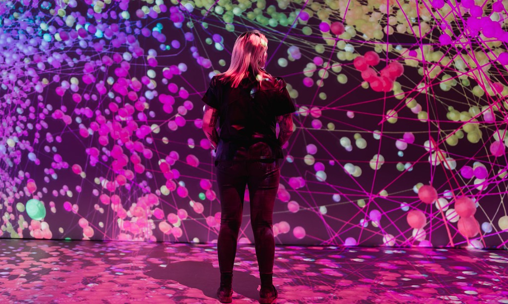 A woman stands by a wall in a digital art gallery and the exhibit on the walls is made up of pink dots and pink lines