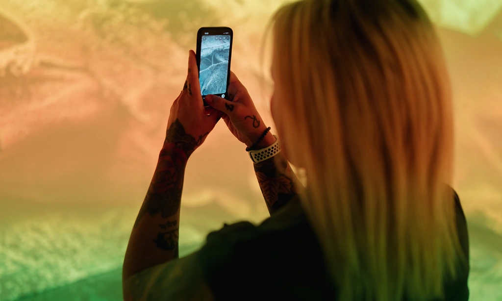 A freelance arts marketer with blonde hair uses an iPhone to take a photo of an exhibition