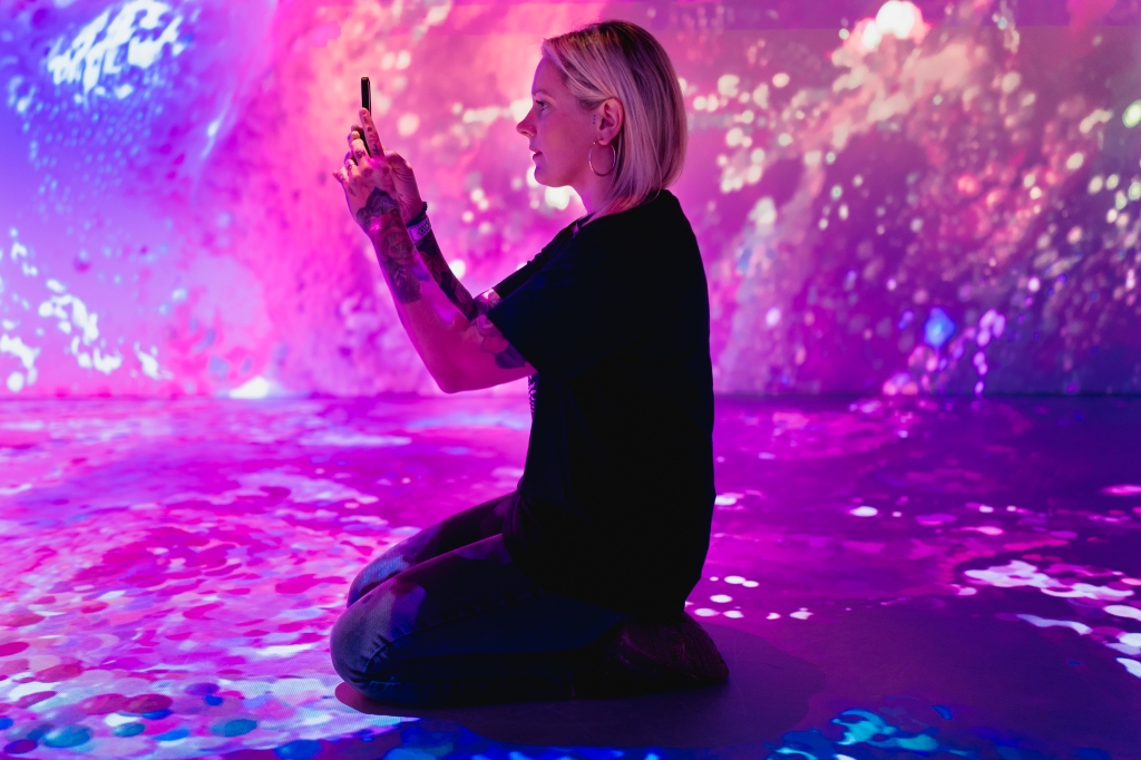 A digital marketing professional takes a photo on an iPhone in the middle of a digital art exhibition in which pink patterns are projected on the walls